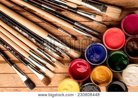 Multicolored Gouache Paints In Open Jars And Artistic Brushes Of Different Sizes On A Light Wooden B