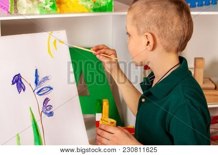 Child painting on easel. Kid boy learn paint by brush in class school. Kindergarten interior on background. Boy is getting ready to become an artist. Quiet environment for children's creativity.
