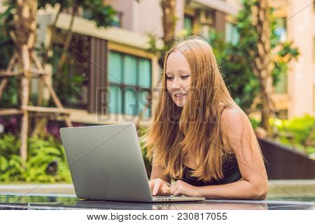 Young Female Freelancer Sitting Near The Pool With Her Laptop In The Hotel Browsing In Her Smartphon
