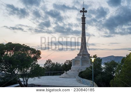 Tower Monument With Christian Cross On Top Against Early Morning Sky In Park, With Street Lights On 