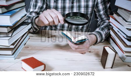 A Man Reading A Miniature Book Among The Big Books In The Library At The Wooden Table.