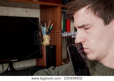 A Thoughtful Guy With A Smartphone At His Mouth, With His Eyes Closed, Sits At The Desk