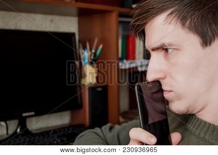 A Thoughtful Guy With A Smartphone At His Mouth, Sits At His Desk, On A Black Monitor Background