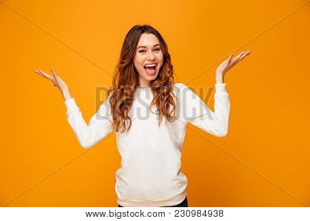 Happy brunette woman in sweater shrugs her shoulders and looking at the camera with open mouth over yellow background
