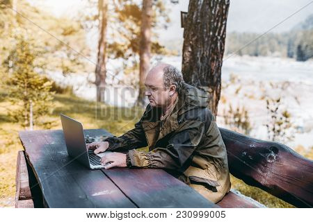 An Adult Partly Bald Game Warden In The Overalls Of Marsh Color Is Sitting Outdoor On The Bench At T
