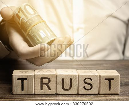 Wooden Blocks With The Word Trust And Money In The Hands Of A Businessman. Trust Relationships Betwe