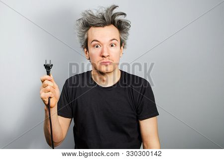 Unshaven Guy With Long Hair Holds An Electric Plug With A Cable In His Hand On A Gray Background