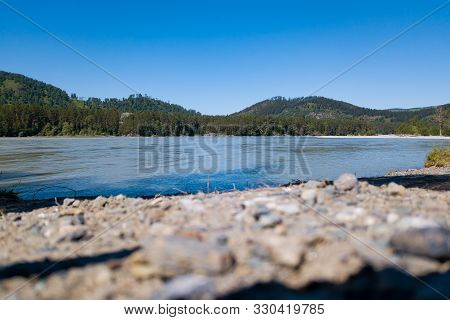 The Shore Of A Mountain River With Stones And Rubble Opposite Which Is A Green Forest With Trees Und