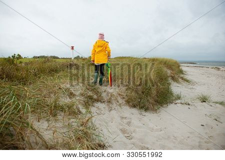 Cheerful Little Girl Staying On Beach With Colorful Ambrella On Baltic Sea At Windy Rainy Weather