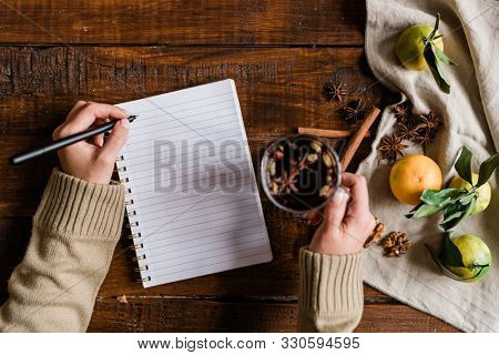 Top view of girl hands holding glass of mulled wine and pen over blank page of notebook while sitting by wooden table