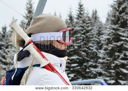 Young, Beautiful Girl In White Ski Suit, With Grey Cap, Googgles And Ski Sticks Using Cable Car, Ski