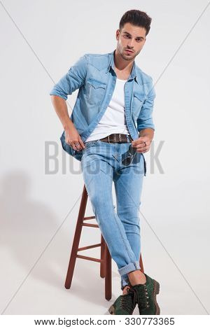 Tough casual man holding his sunglasses adn adjusting hsi jacket while wearing jeans and jacket, leaning on a stool light gray background