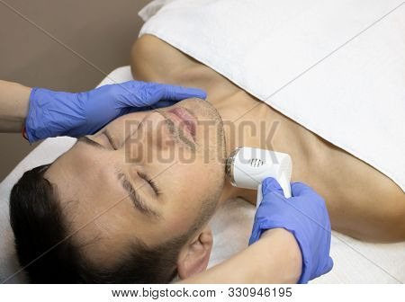 Young Man Laying On A Facial Therapy Couch Covered With The White Towel With Opened Eyes. Facial The