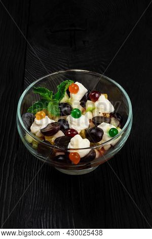 Salad from grapes apples pears kiwi oranges with mascarpone chease and cream. Healthy fresh fruit summer salad in glass bowl on black wooden background with copy space.