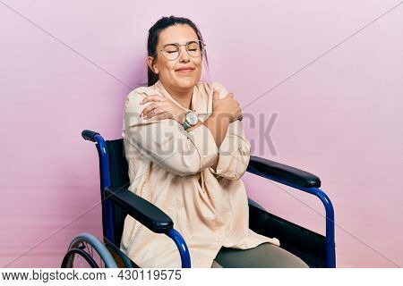 Young hispanic woman sitting on wheelchair hugging oneself happy and positive, smiling confident. self love and self care 
