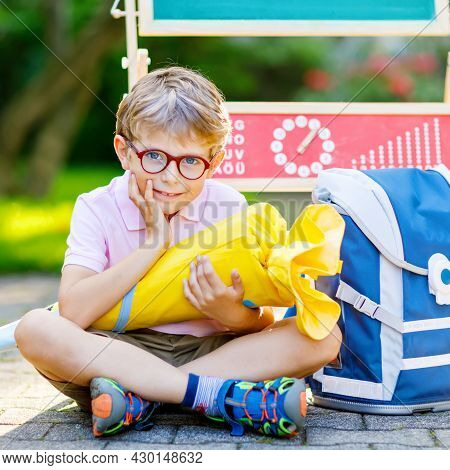 Happy Little Kid Boy With Glasses Sitting By Desk And Backpack Or Satchel. Schoolkid With Traditiona