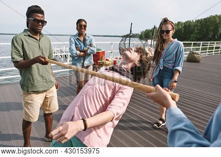 Group of joyful young people playing limbo game on pier by the lake when enjoying small party