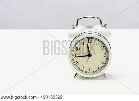 White Round Alarm Clock Stands On The Table, The Time Is Fifteen Minutes To Twelve