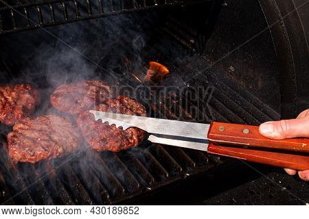 Roasted Meat Bbq Burger Patty For Hamburger In Metallic Tongs Utensils Over Flaming Grill Barbecue W