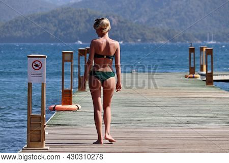 Hisaronu, Turkey - August 2021: Blonde Girl In Green Bikini Walking By Wooden Pier On Sea And Mounta