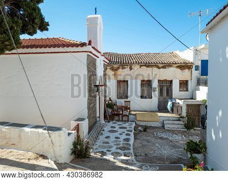 Old rusty traditional roof tiled house next to Cyclades architecture Greek Orthodox Christian Chapel at Kythnos island, Messaria town Greece. Asphalt empty yard with paint pictures on, chairs, plants.