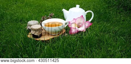 A Cup Of Tea And A Teapot On A Wooden Stand With A Jar Of Honey And Pink Flowers Among The Grass