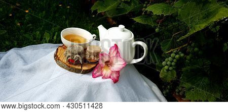 A Cup Of Tea And A Teapot On A Wooden Stand With Jars Of Honey And Pink Flowers Among Grapes