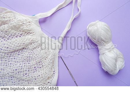Eco Bag In The Knitting Process. White Grid Bag, Crochet Hook And Remaining Yarn On A Purple Backgro