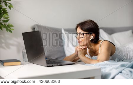 Adorable Woman In Wireless Earhones And Eyeglsees Resting Duirng Morning Time On Bed And Surfing Int