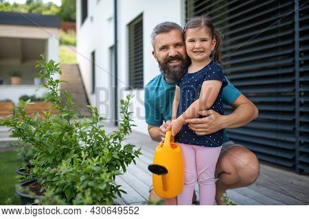 Father And Small Daughter With Watering Can Outdoors In Tha Backyard, Looking At Camera.