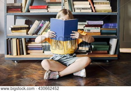 Little Girl Sitting With Open Fairy Tale In Front Of Her Face On Wooden Parquet Against Bookshelf Fu