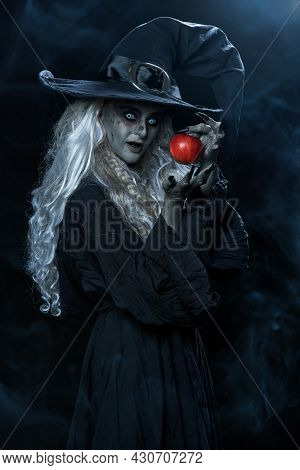 Halloween. Scary green old witch holds out a red apple. Woodland background, evil black magic.