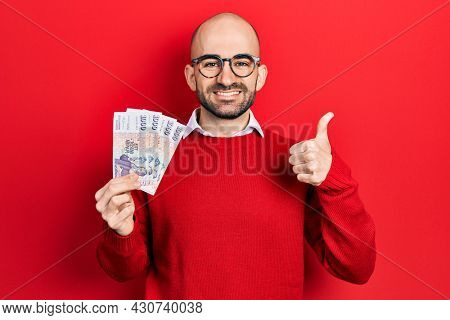 Young bald man holding icelandic krona banknotes smiling happy and positive, thumb up doing excellent and approval sign 