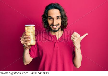 Young hispanic man holding bottle with boiled potatoes pointing thumb up to the side smiling happy with open mouth 