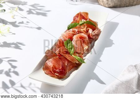 Italian antipasti on white table. Meat starter - sliced meat with basil on white dish. Sunshine and shadow on white table. Traditional italian first course include prosciutto, bresaola and parma ham