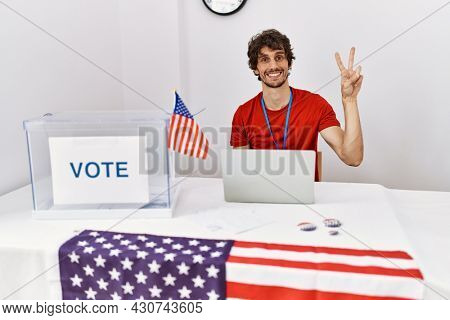 Young hispanic man at political election sitting by ballot showing and pointing up with fingers number two while smiling confident and happy. 