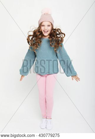 funny child girl with curly haor dressed blue sweater and winter hat jumping on white background