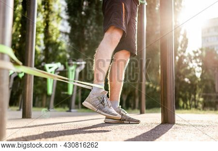 Strong man guy training with elastic rubber band at the stadium outdoors. Adult male person during workout for legs with additional sport equipmant. Sportsman exercising outside