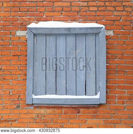 Boarded Up Window With Gray Painted Planks Against A Bright Red Brick Wall In Winter Covered With Sn