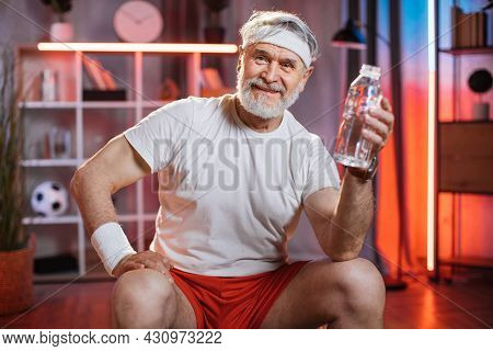 Senior Caucasian Man In Sport Clothes And Headband Sitting On Fitball With Bottle Of Water In Hand. 