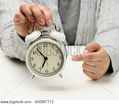 Female Hand Holds A White Metal Alarm Clock On A White Table, Time Five Minutes To Seven In The Morn