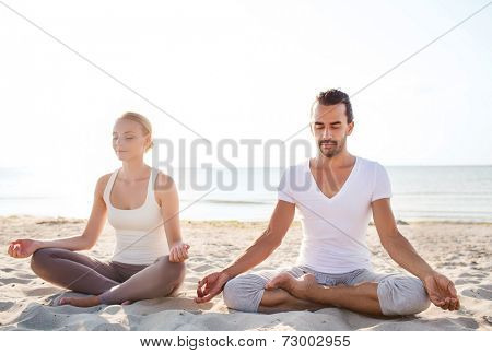 fitness, sport, friendship and lifestyle concept - smiling couple making yoga exercises sitting on sand outdoors