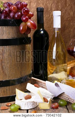 Supper consisting of Camembert and Brie cheese, wine and grapes on napkin in basket and wine barrel on wooden table on brown background