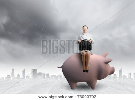 Young businesswoman with suitcase in hand sitting on piggybank