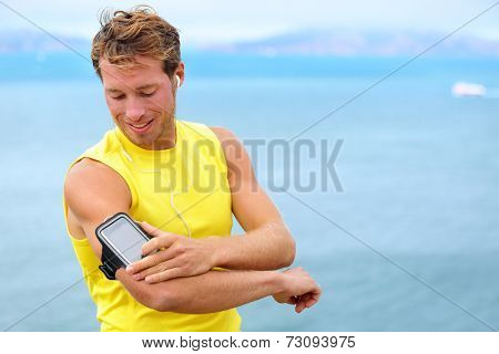 Running training music on smartphone app. Runner man listening to music adjusting settings on armband for smart phone. Fit male fitness model working outdoor by water.
