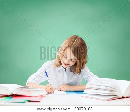 education, people, children and school concept - happy student girl sitting at table with books and writing in notebook over green chalk board background