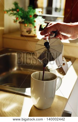 closeup of a young caucasian man serving coffee in a white mug from a moka pot in the kitchen