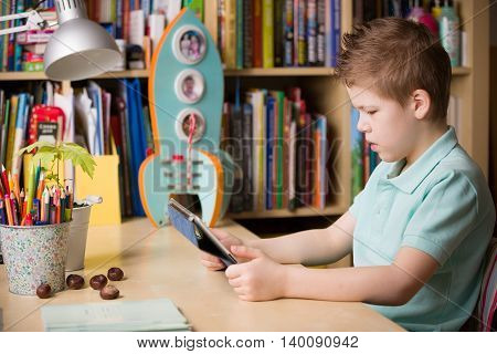 School kid boy sitting at his table and watching tablet pc at home indoors. Child with tablet computer in his room. Studying with tablet computer.