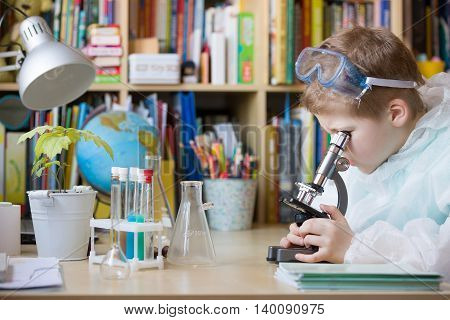 Cute elementary school boy looking into microscope at his desk at home. Young scientist making experiments in his home laboratory. Indoors. Child and science.
