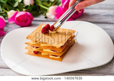 Shortcake with berries. Hand holding tongs. Chef prepares raspberry millefeuille. Layered dessert with sweet filling.